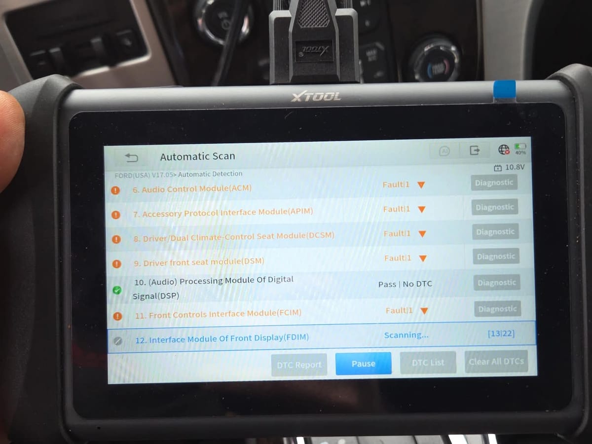 Diagnostic scan tool held beside a vehicle part way through an automatic scan, listing faults module by module